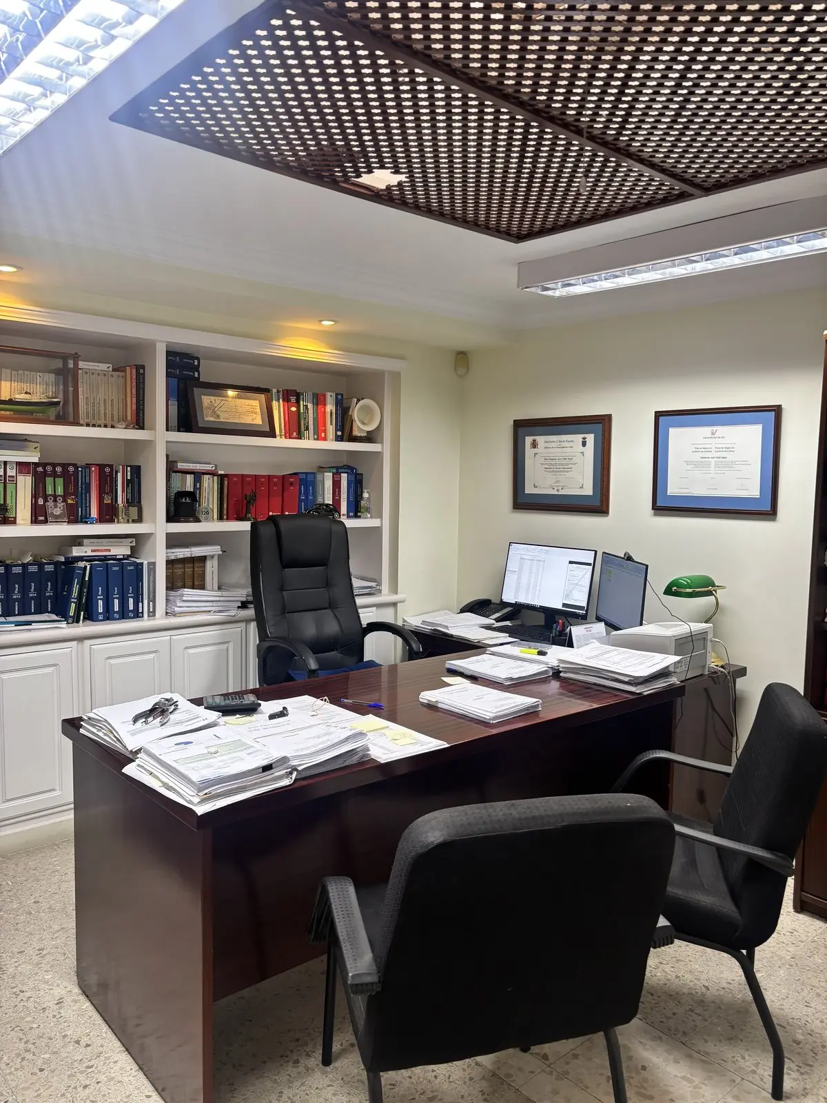 Despacho con la mesa de reuniones, la biblioteca y las titulaciones colegiales enmarcadas
